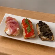 Tris Bruschette — Three bruschette (toasted bread): cherry tomatoes with oregano — cured ham with stracciatella — grilled aubergine with smoked provola